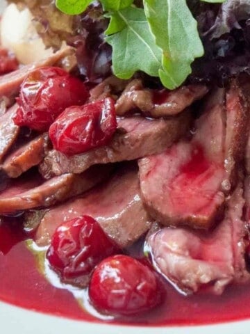 Sliced duck breast meat covered in cherries and red sauce on a white plate with salad on the side.