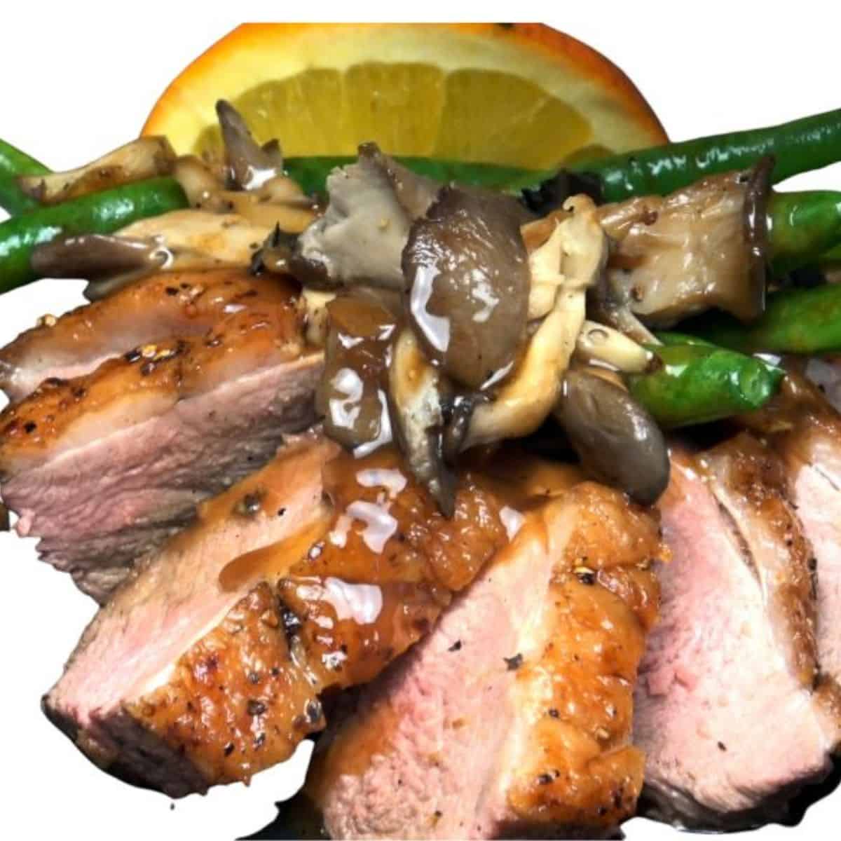 Pink slices of duck breast with golden brown skin with vegetables and lemon covered in a slightly orange sauce.