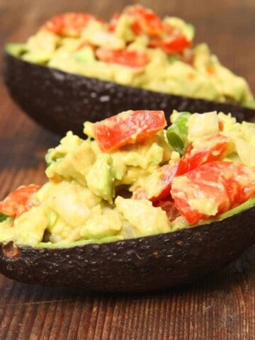 Two dark green avocado shells stuffed with red tomatoes and other ingredients covered in cream colored sauce on a wood surface