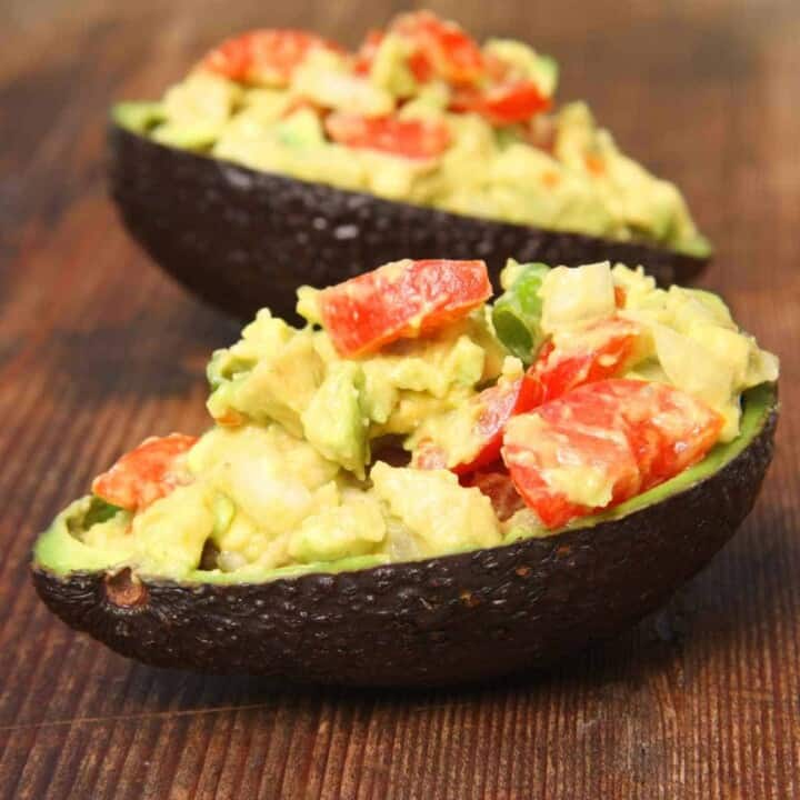 Two dark green avocado shells stuffed with red tomatoes and other ingredients covered in cream colored sauce on a wood surface