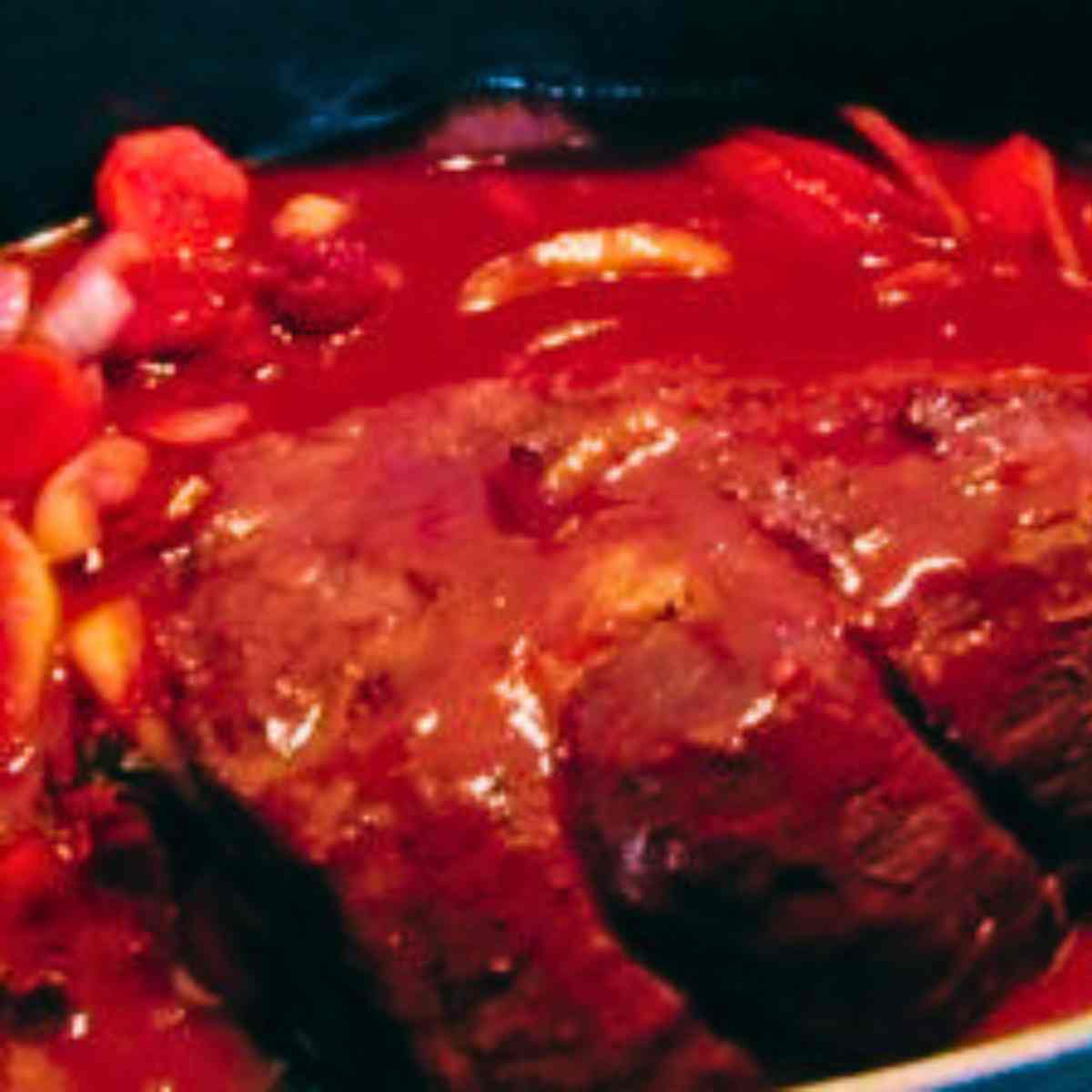 Large slab of red meat, carrots, celery, and onions covered in red tomato marinade.