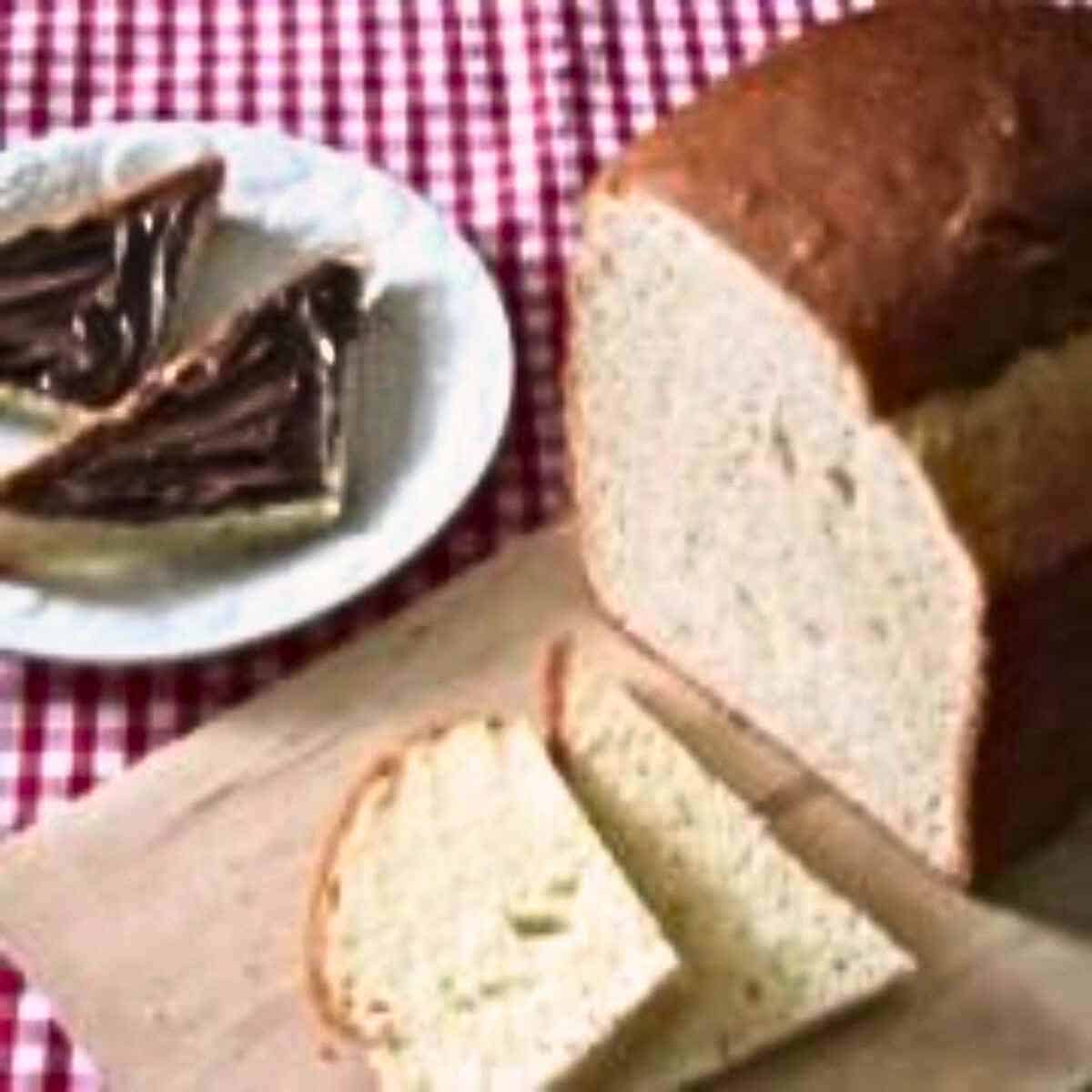 A loaf of white bread with brown crust on a wooden cutting board with slices and a white side plate with sliced spread with chocolate spread all on a red gingham table cloth.