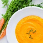 white bowl of creamy, orange, carrot soup with leafy garnish and fresh carrots on side with greenery.