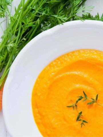 white bowl of creamy, orange, carrot soup with leafy garnish and fresh carrots on side with greenery.