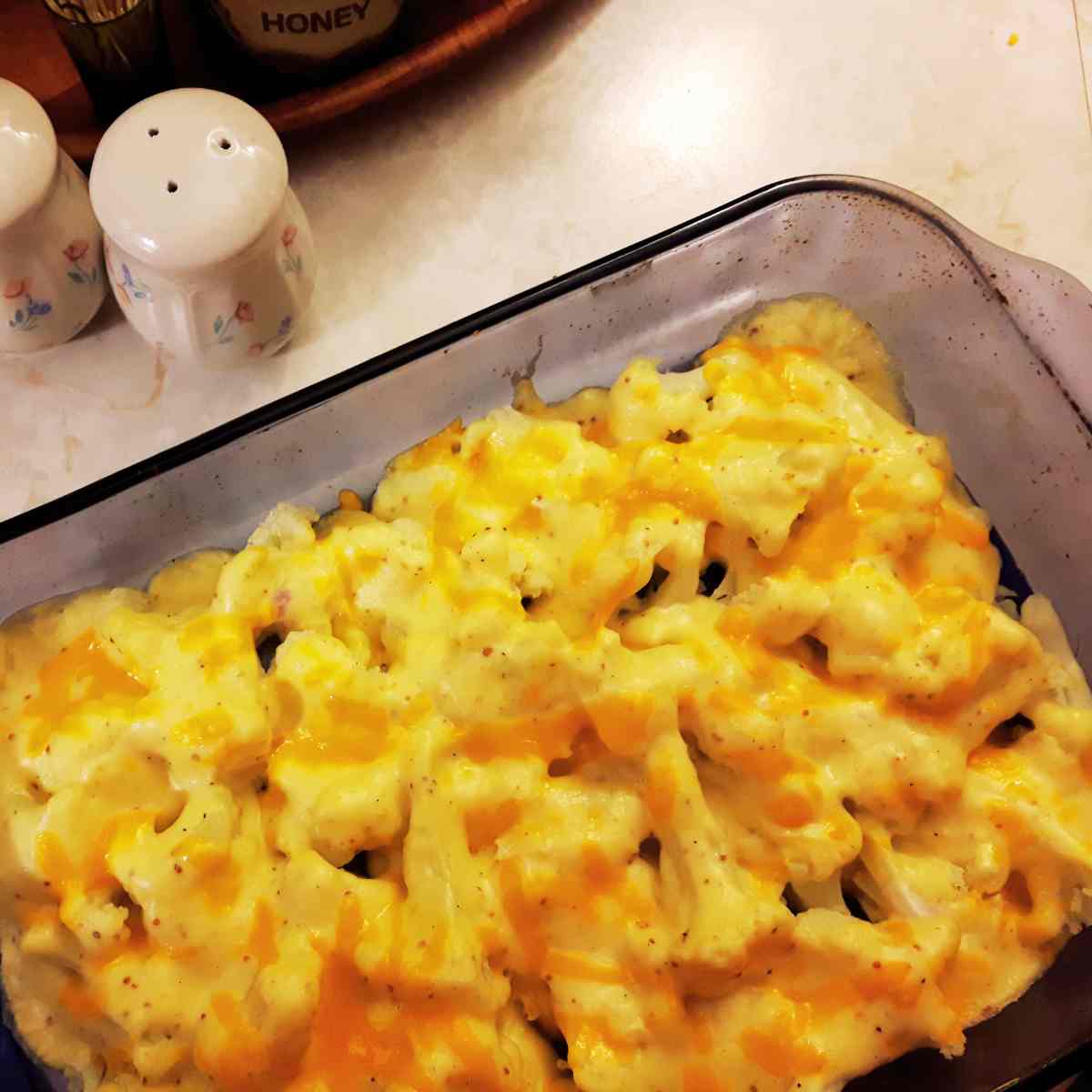 Dark glass oven proof dish with cauliflower pieces covered in a sauce with yellow cheese on top with salt and pepper on side.