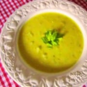 A white ornate soup bowl on a red gingham table cloth filled with creamy light green celery soup topped with green leaves.