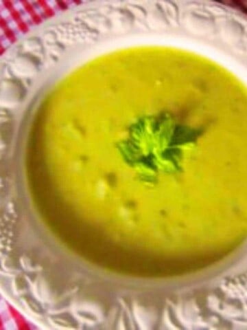 A white ornate soup bowl on a red gingham table cloth filled with creamy light green celery soup topped with green leaves.