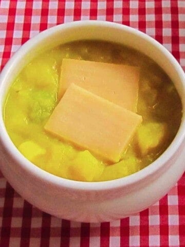 A white soup bowl filled with chunky, creamy potato soup with 2 slabs of orange cheese on top on a red gingham tablecloth.