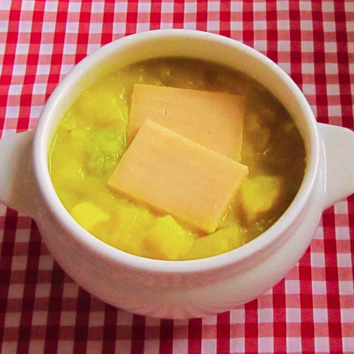 A white soup bowl filled with chunky, creamy potato soup with 2 slabs of orange cheese on top on a red gingham tablecloth.