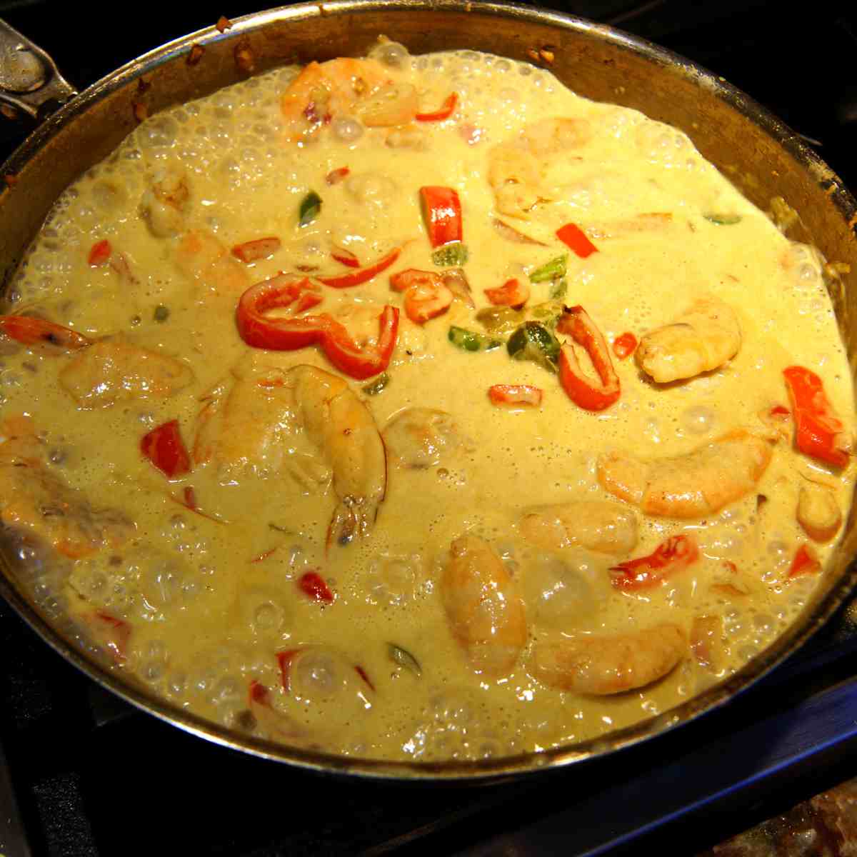 A large dark colored frying pan full of yellow curry dish with king shrimp, red peppers and other vegetables and chunks of meat cooking and bubbling.