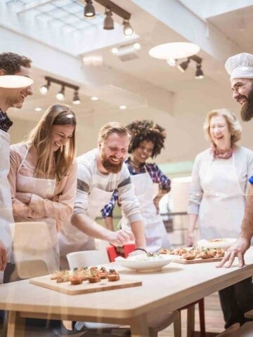 A group of cooking students with a chef staning around a table with food on it.