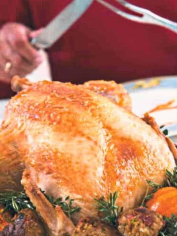 A large roasted turkey on a plate with vegetables around it and a man holding a knife and fork ready to carve the bird.