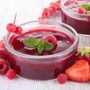 Two clear glass bowls filled with red thick juice, decorated with a raspberry and mint leaf and surrounded with strawberries, raspberries, and round red berries.