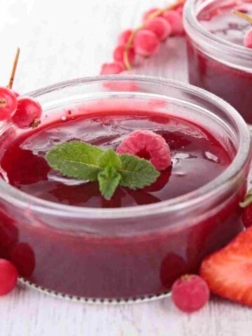 Two clear glass bowls filled with red thick juice, decorated with a raspberry and mint leaf and surrounded with strawberries, raspberries, and round red berries.