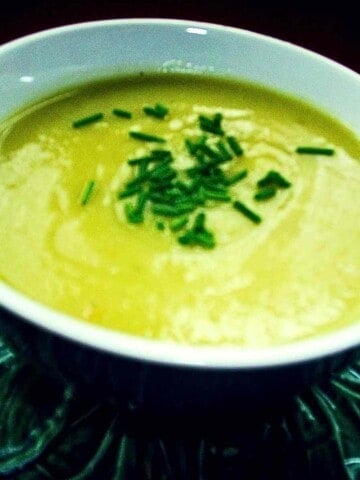 A white soup bowl filled with a creamy light green soup of blended leeks and potatoes sprinkled with dark green chives.