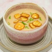 Brown soup bowl on a saucer filled with creamy parsnip soup topped with toasted parsnip rounds and flakes of parsley.