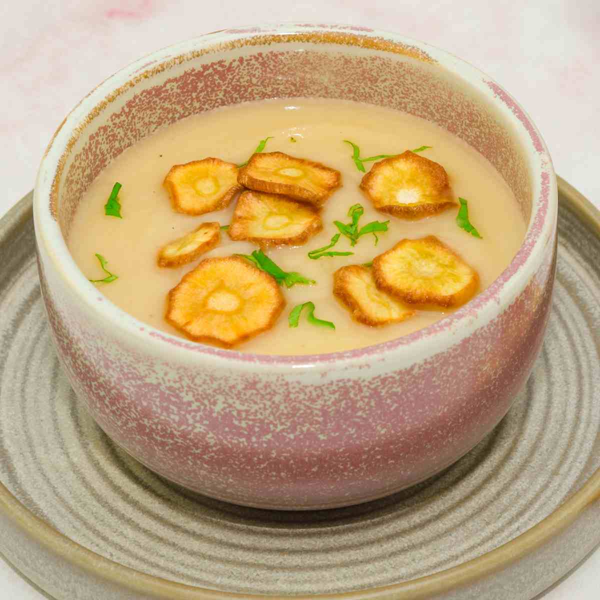 Brown soup bowl on a saucer filled with creamy parsnip soup topped with toasted parsnip rounds and flakes of parsley.