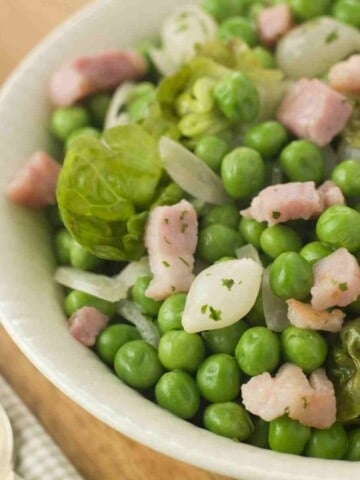 A white bowl filled with green peas with bits of lettuce, onion, and pale pink bacon.