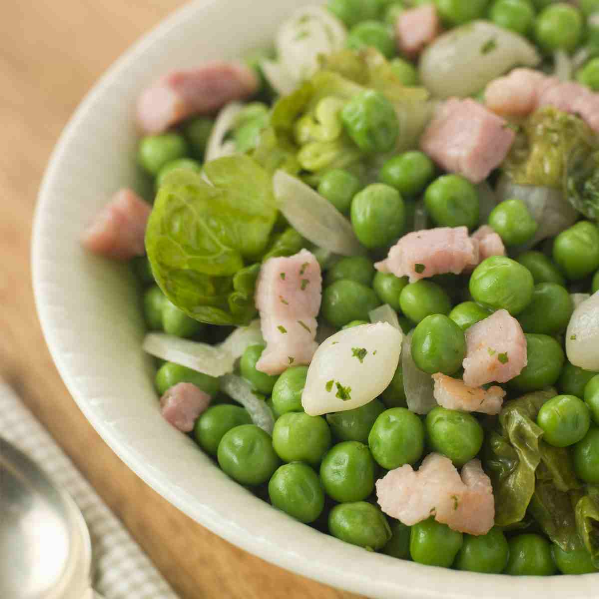 A white bowl filled with green peas with bits of lettuce, onion, and pale pink bacon.