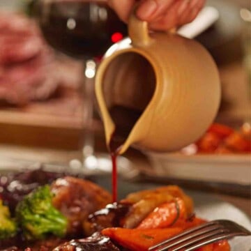 Cream colored gravy boat being poured onto beef with a red wine jus sauce.