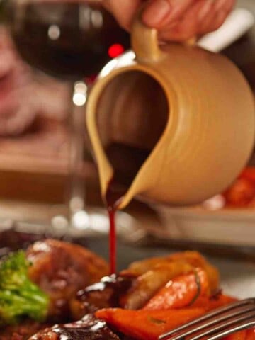 Cream colored gravy boat being poured onto beef with a red wine jus sauce.