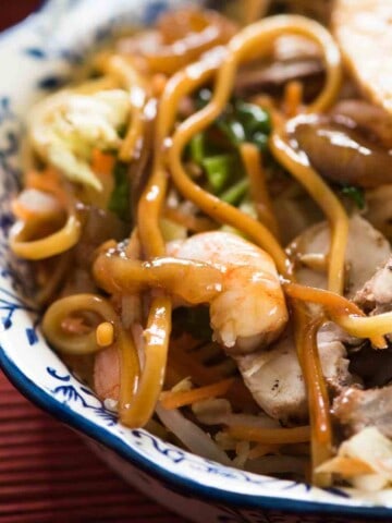 A white and blue bowl with large flat egg noodles, green vegetable, pink shrimp, brown sliced meat, all covered in a light brown sauce.