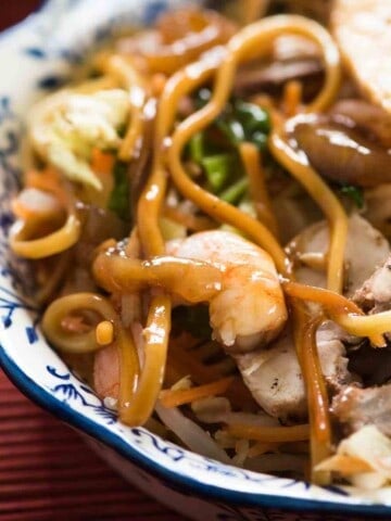 A white and blue bowl with large flat egg noodles, green vegetable, pink shrimp, brown sliced meat, all covered in a light brown sauce.