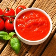 A white bowl filled with red pureed tomatoes with 5 small red tomatoes on a stem with basil all on a wood background.