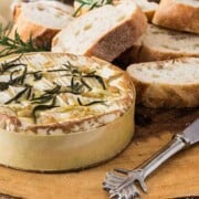 A round white camembert cheese sprinkled with rosemary and slices of white, brown crusted baguette bread beside it.