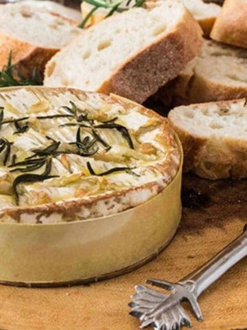 A round white camembert cheese sprinkled with rosemary and slices of white, brown crusted baguette bread beside it.