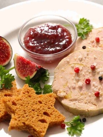 A white dinner plate with two round slabs of beige slices of duck foie gras with red berries on top, star-shaped gingerbread, slices of figs, and a bowl of red fig jam.