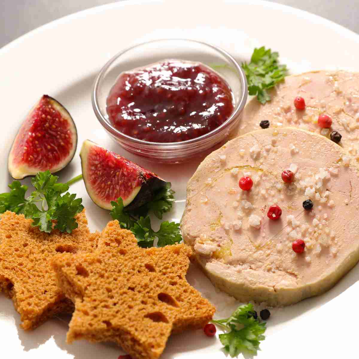 A white dinner plate with two round slabs of beige slices of duck foie gras with red berries on top, star-shaped gingerbread, slices of figs, and a bowl of red fig jam.