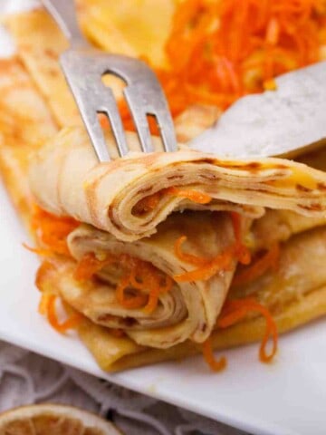 A white plate full of thin, golden, rolled pancakes called crepes with grated orange rind.