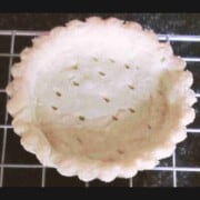 An off-white pie shell with scalloped edges and steam release holes across the bottom cooling on a wire rack.