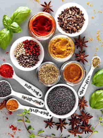 Small bowls and spoons of various colorful seasonings with star anise, green basil leaves and spices scattered around.