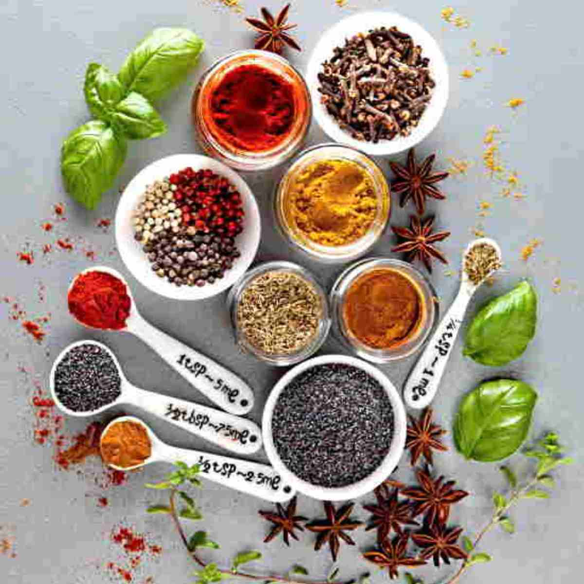 Small bowls and spoons of various colorful seasonings with star anise, green basil leaves and spices scattered around.