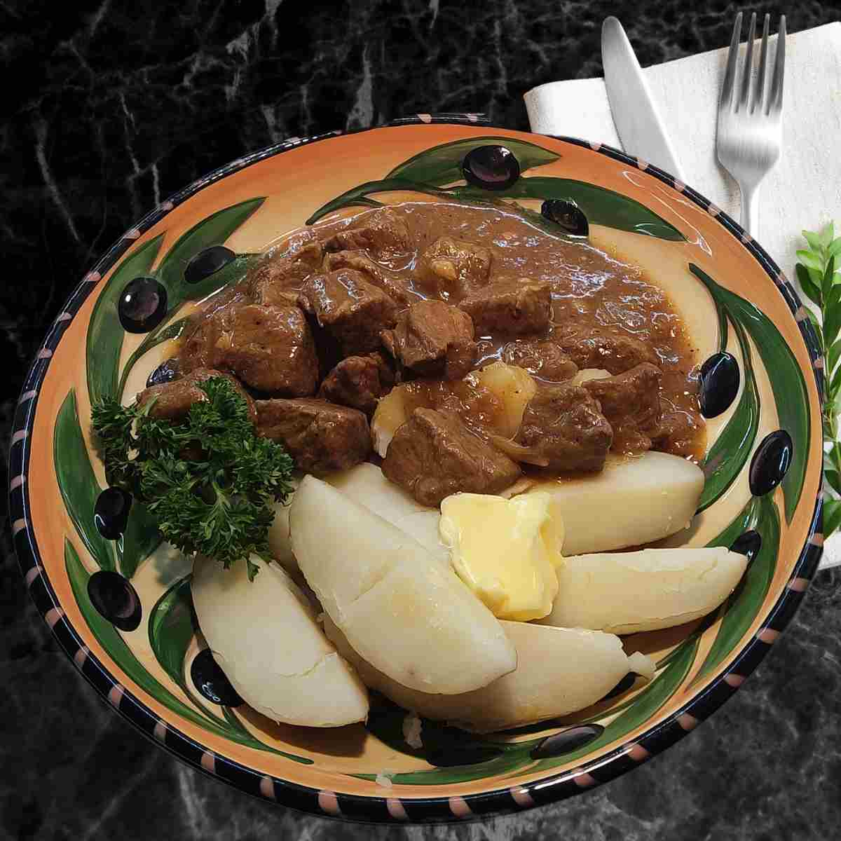 A large shallow bowl with black olives and green leaf pattern with brown beef chunks in sauce beside white potato wedges, pad of yellow butter and green sprig of parsley on side.