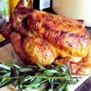 Golden brown roasted whole chicken with a sprig of tarragon on a white cutting board.