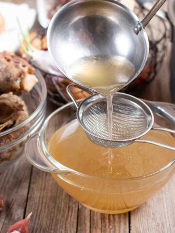 Clear glass bowl of light brown liquid (broth) surrounded by vegetables, bowl of meat bones, and large soup pot.