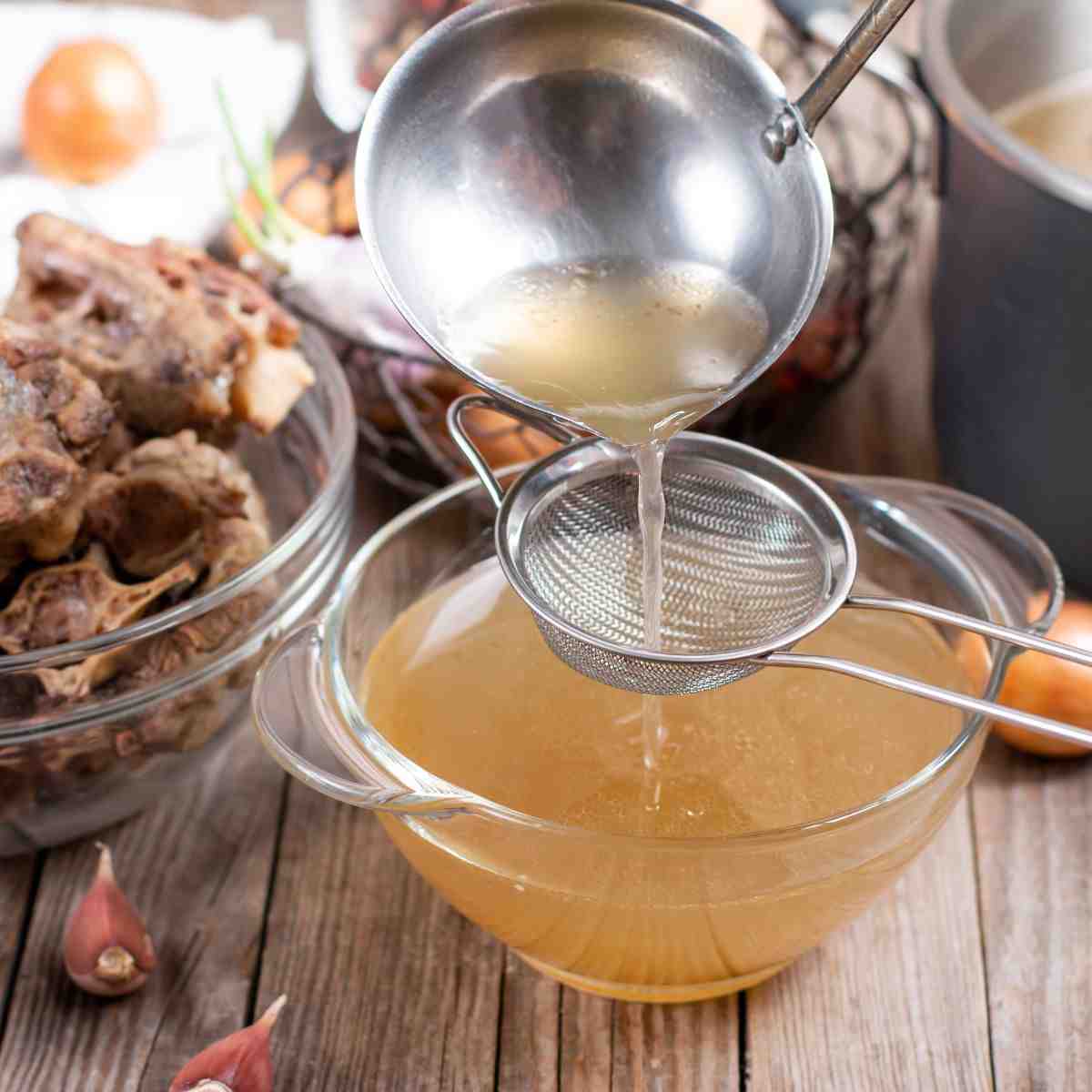 Clear glass bowl of light brown liquid (broth) surrounded by vegetables, bowl of meat bones, and large soup pot.