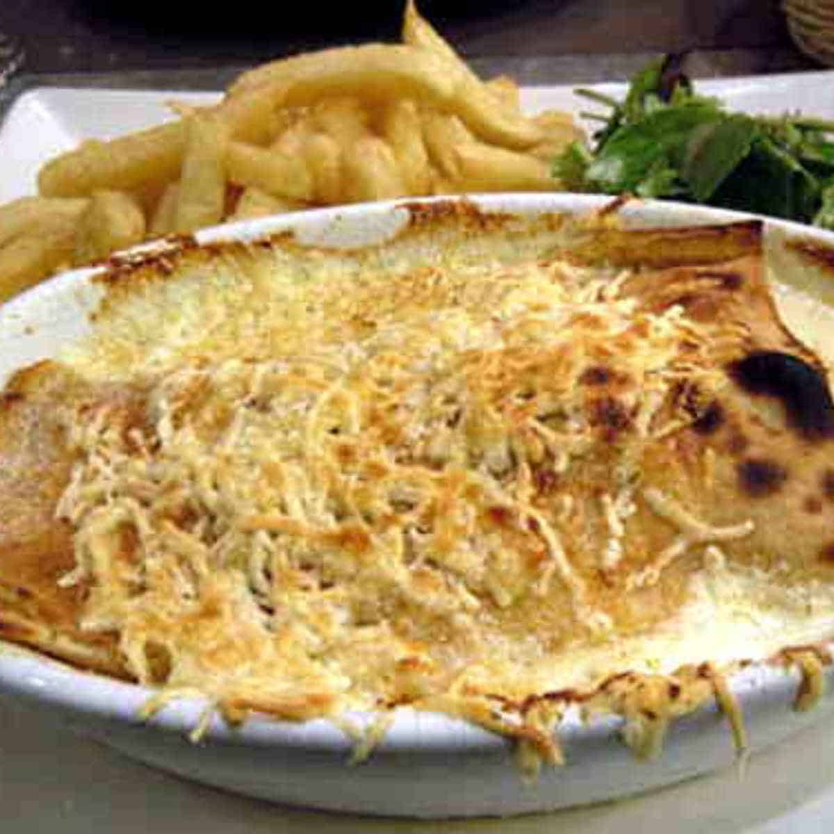 A stuffed crepe with golden brown melted cheese on top with golden french fries and green leaves.