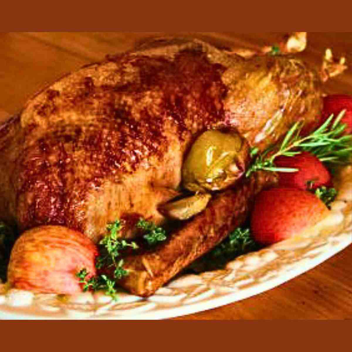 A whole roasted goose, different shades of brown surrounded by red apples, and green herbs.