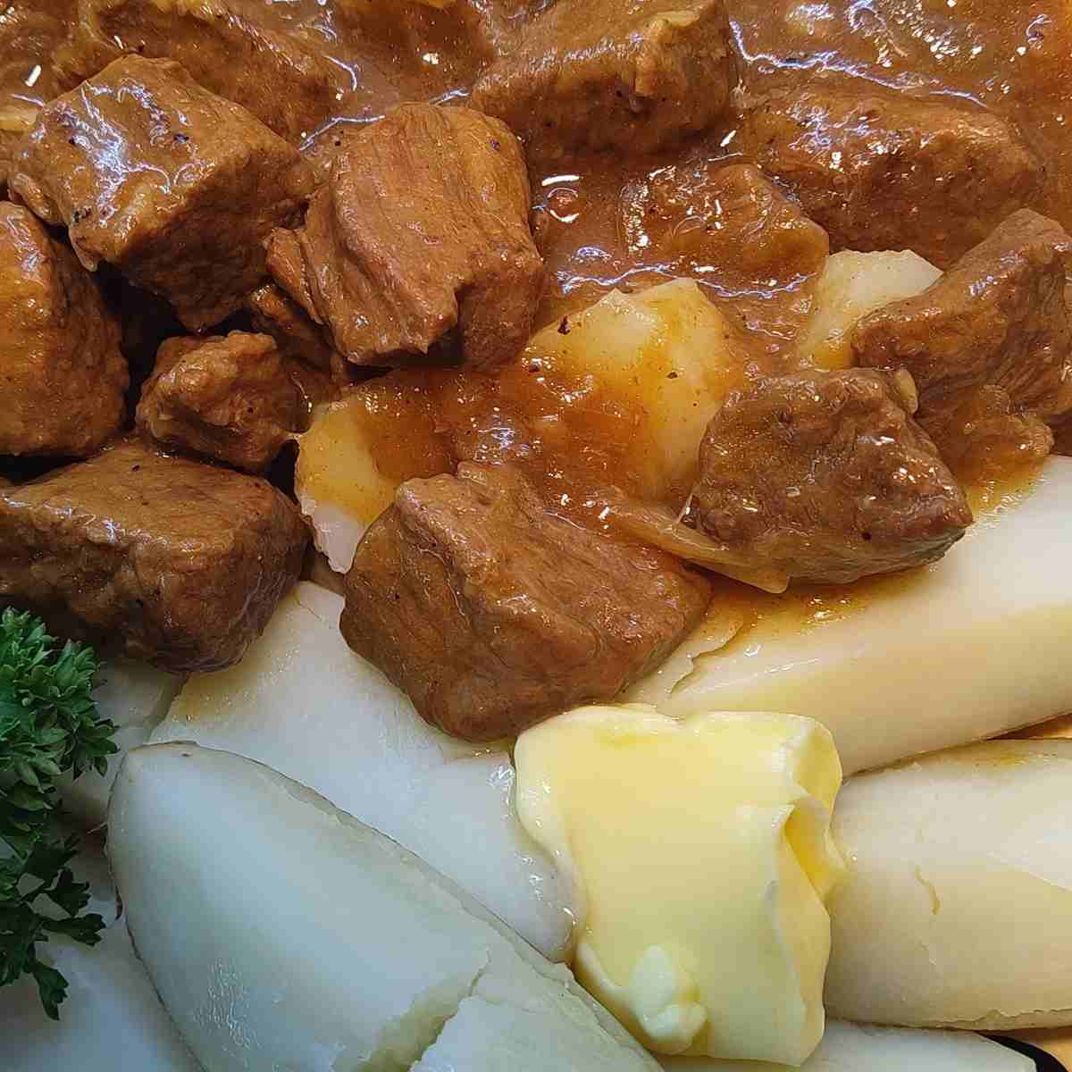 Chunks of beef in brown gravy beside boiled potato wedges with a pad of butter and parsley on the side.