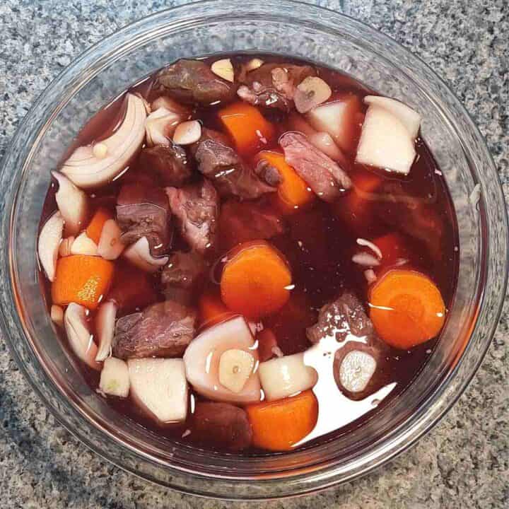 A bowl of redish brown beef chunks, white chopped onion, shallots, garlic, and orange carrots in a red wine marinade.