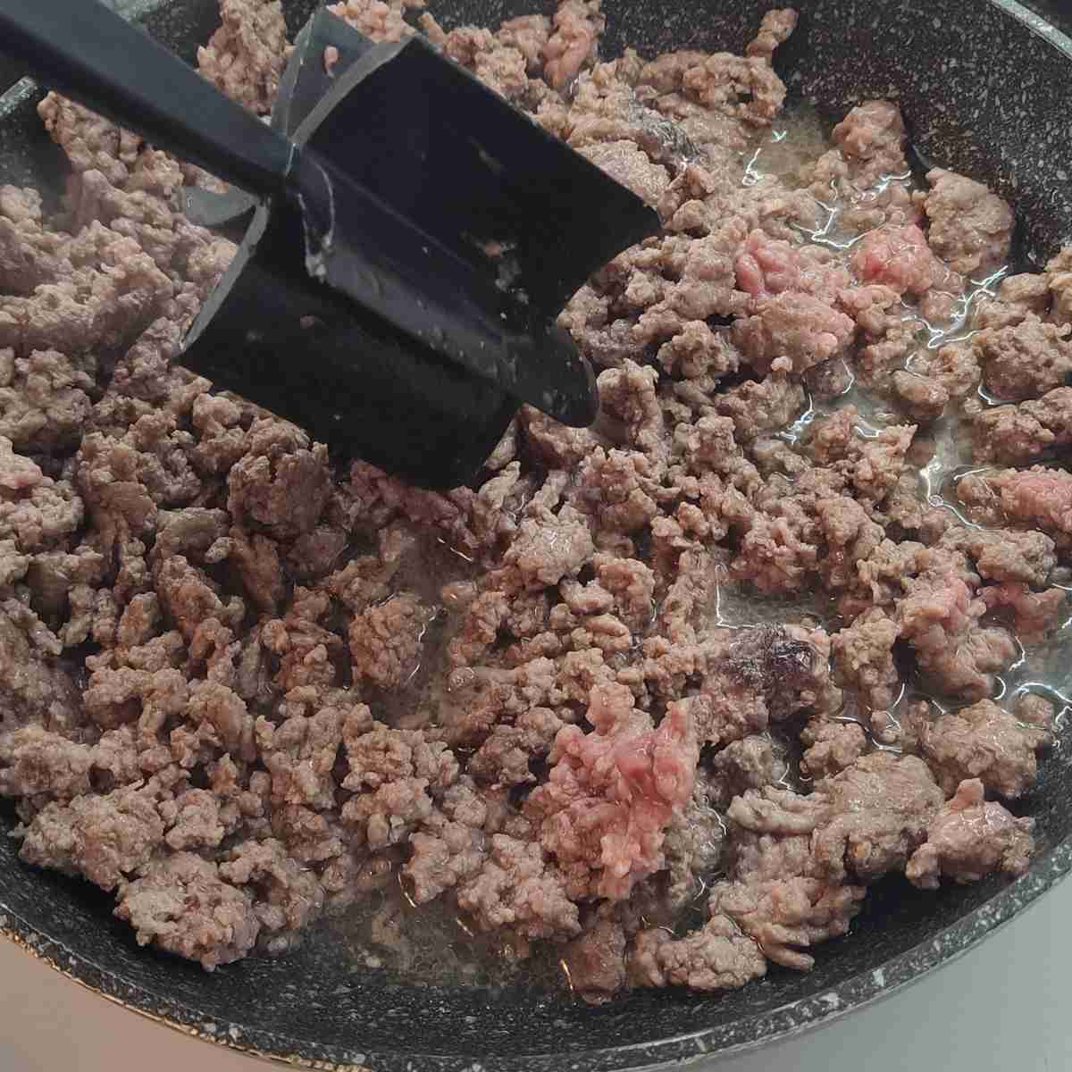 Raw red ground beef cooking to a brown in a black pan with a black mix'n chop cooking tool.