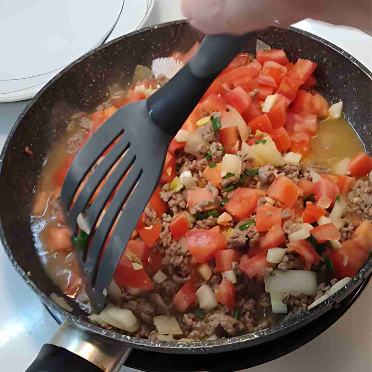Black frying pan full of brown ground beef, red tomatoes, and other white and yellow vegetables.