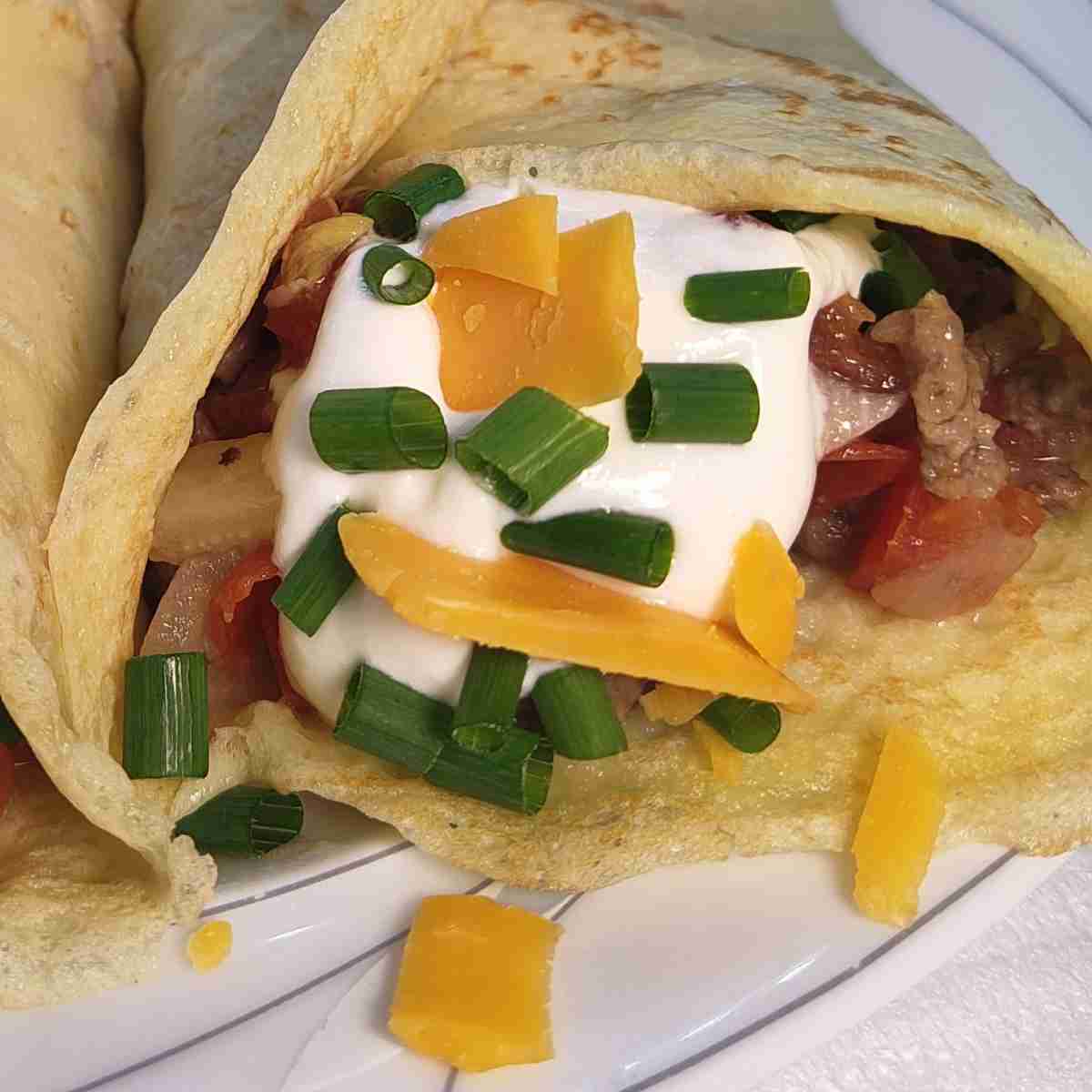 A beige crepe rolled around a filling of red tomatoes, brown meat, white sauce on top, sprinkled with green chives, and grated orange cheese.