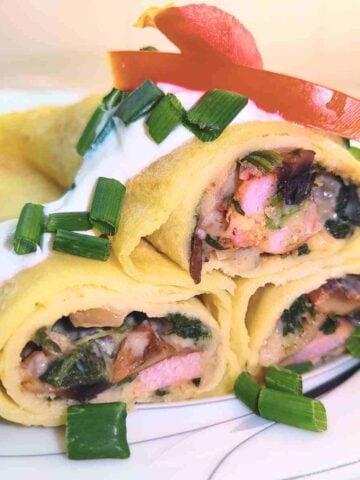 Three stuffed crepes piled on top of one another, garnished with red tomato and green chives on white sour cream.