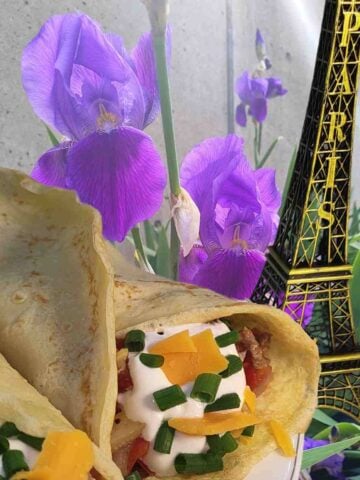 Beige rolled crepe with brown meat, red tomato, green chives, and orange cheese filling with a gold Eiffel tower icon and the French national flower, purple iris.