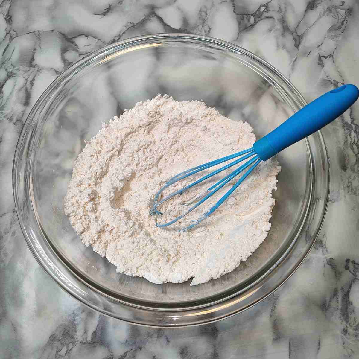 Dry ingredients in clear glass bowl with blue whisk.